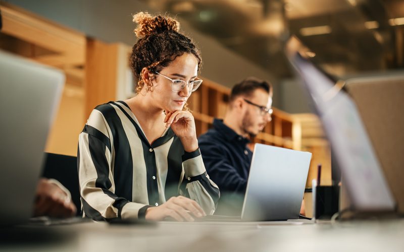 Portrait of Enthusiastic Hispanic Young Woman Working on Computer in a Modern Bright Office. Confident Human Resources Agent Smiling Happily While Collaborating Online with Colleagues.